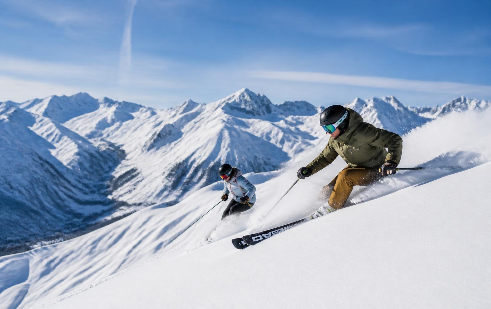 Two skiers carving through powder snow against an Alpine panorama